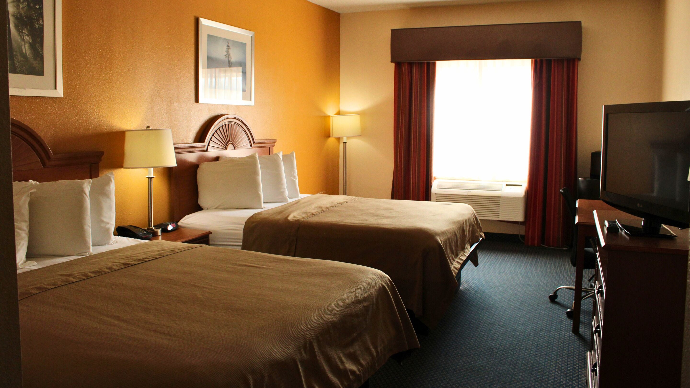 A hotel room with two beds, a desk with a television, a lamp, and a window with curtains.