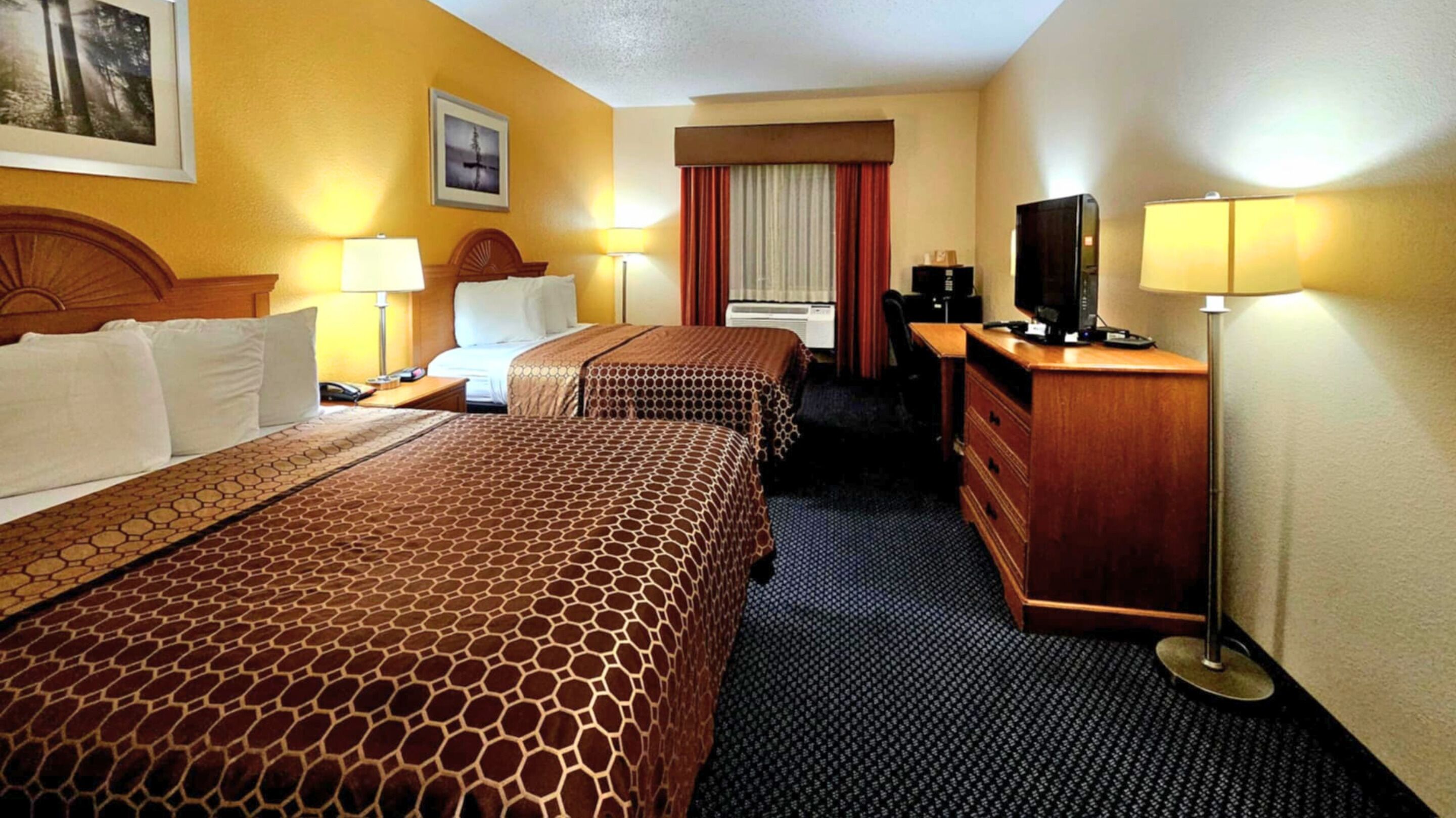 Una habitación de hotel con dos camas, un escritorio con televisor, un aparador y una ventana con cortinas.