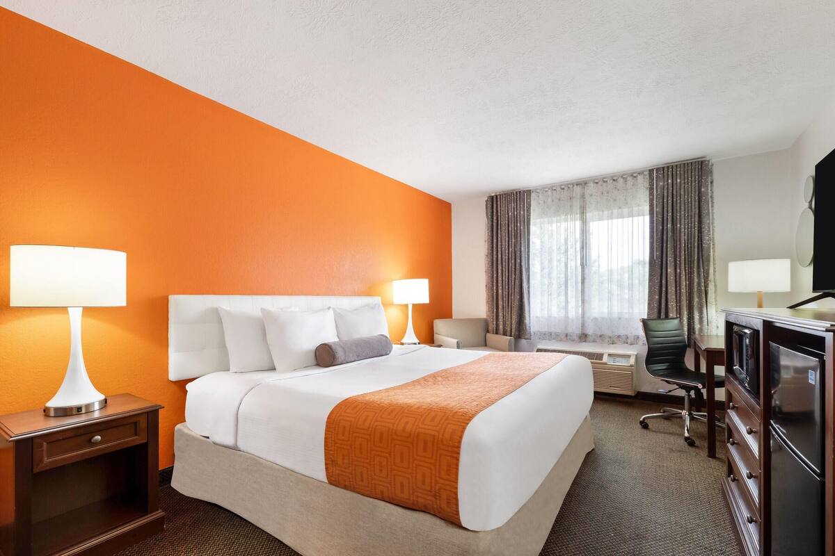Ein Hotelzimmer mit einem großen Bett, zwei Nachttischen mit Lampen, einem Schreibtisch, einem Sessel, einem Fernseher und einem Fenster mit Vorhängen.