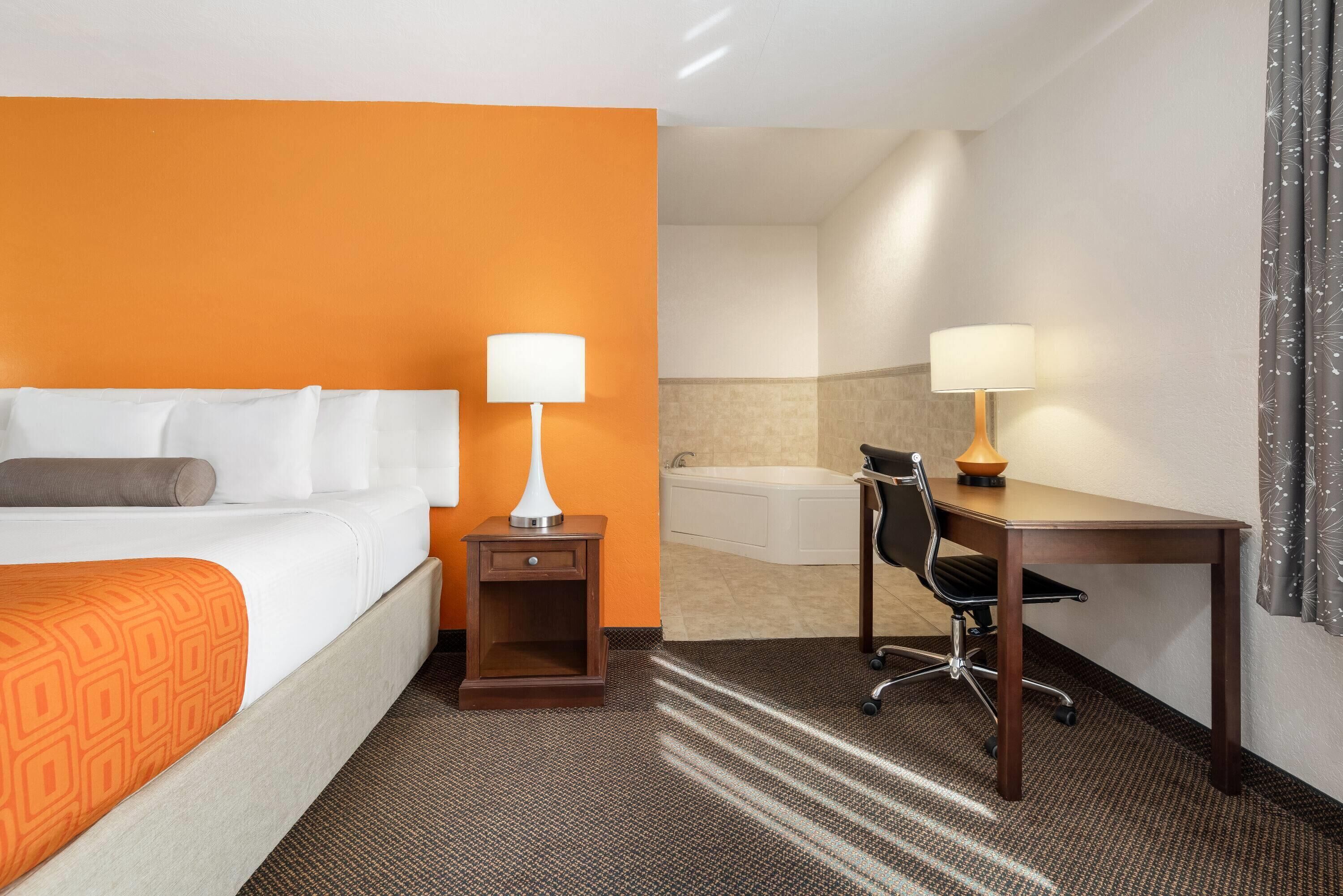 Une chambre d’hôtel comprenant un lit, un bureau, une chaise, une lampe, une table de chevet et une salle de bain avec une baignoire.