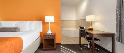 A hotel room with a bed, a desk, a chair, a lamp, a nightstand, and a bathroom with a bathtub.