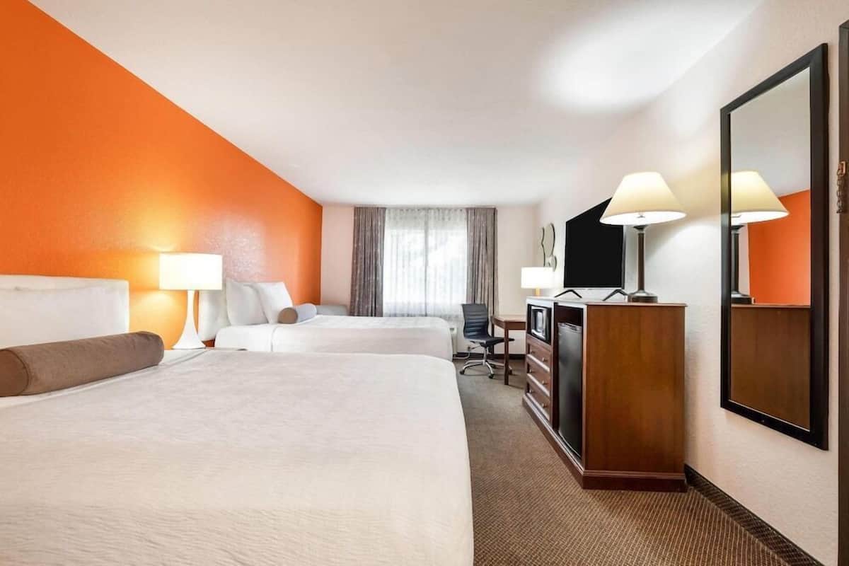 Ein Hotelzimmer mit zwei Betten, einem Schreibtisch, einem Fernseher, einem Spiegel und einem Fenster mit Vorhängen.