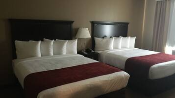 Habitación de hotel con dos camas, cada una con ropa de cama blanca y un detalle rojo. Entre las camas hay una mesita de noche con una lámpara.