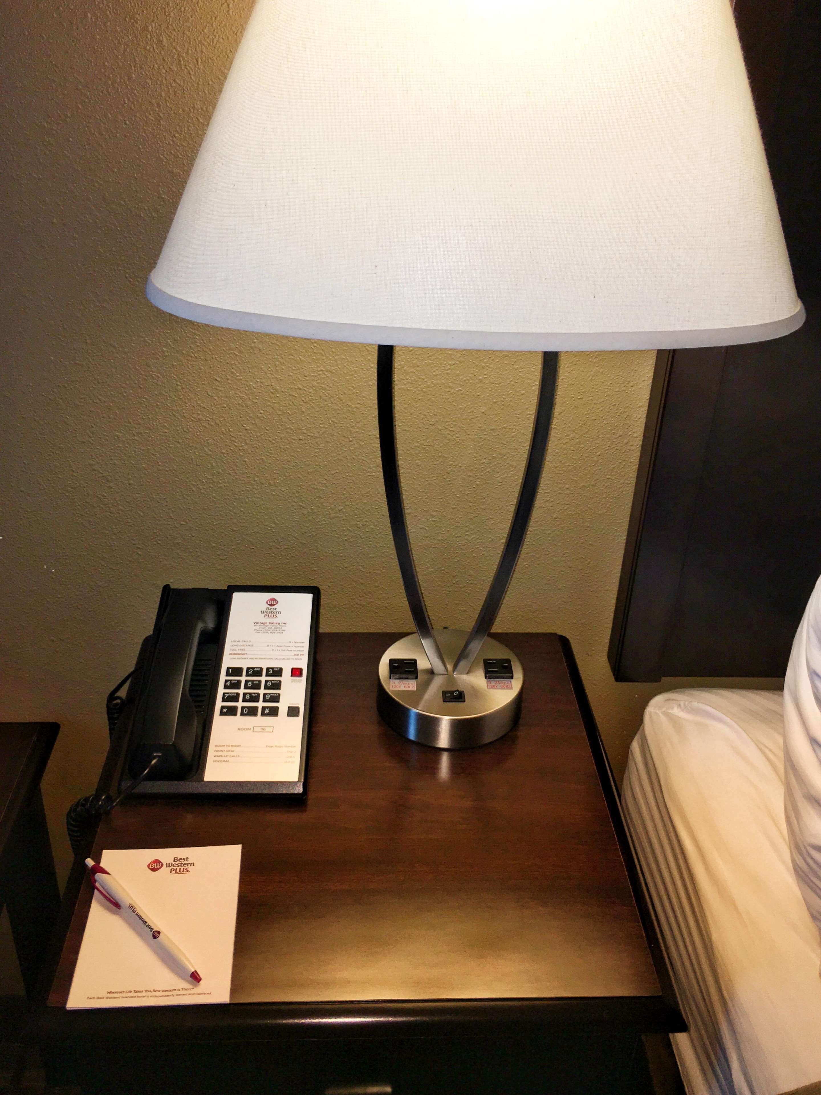 Hotelkamer met nachtkastje, lamp, telefoon en pen op notitieboekje.