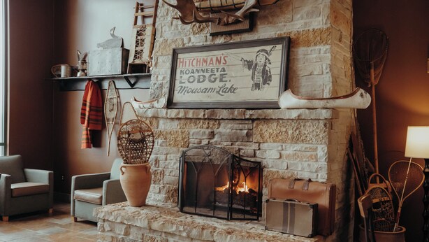 A stone fireplace with a framed sign above it, two armchairs, a basket, and a bowl of fruit on the floor. - Wildwood Lodge (Pewaukee)