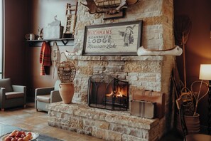 Una chimenea de piedra con un cartel enmarcado encima, dos sillones, una cesta y un bol de fruta en el suelo.