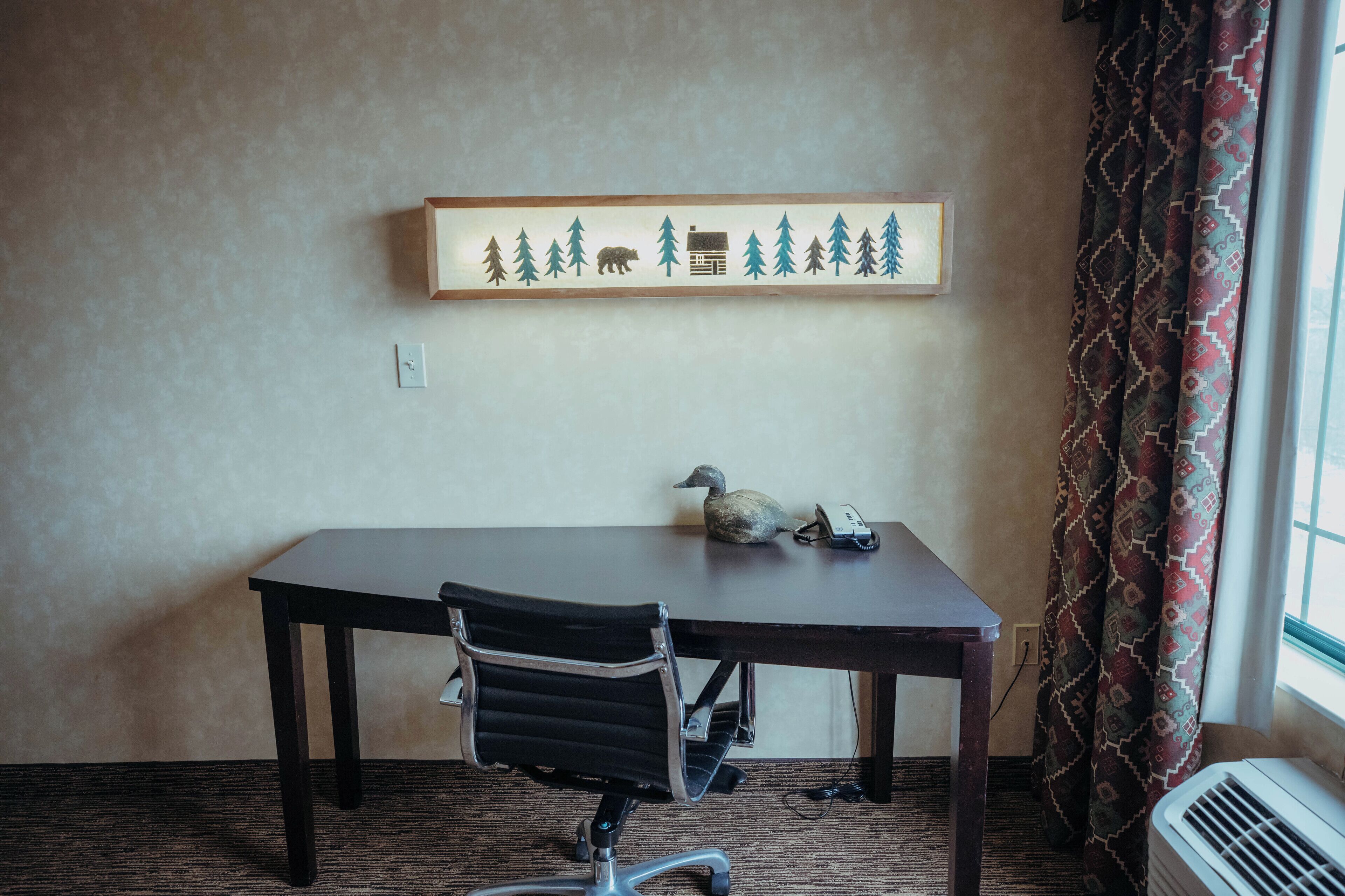 Une pièce avec une table en bois foncé, un fauteuil de bureau noir, un tableau encadré représentant un ours et des arbres accroché au mur, un téléphone et un rideau à motifs.
