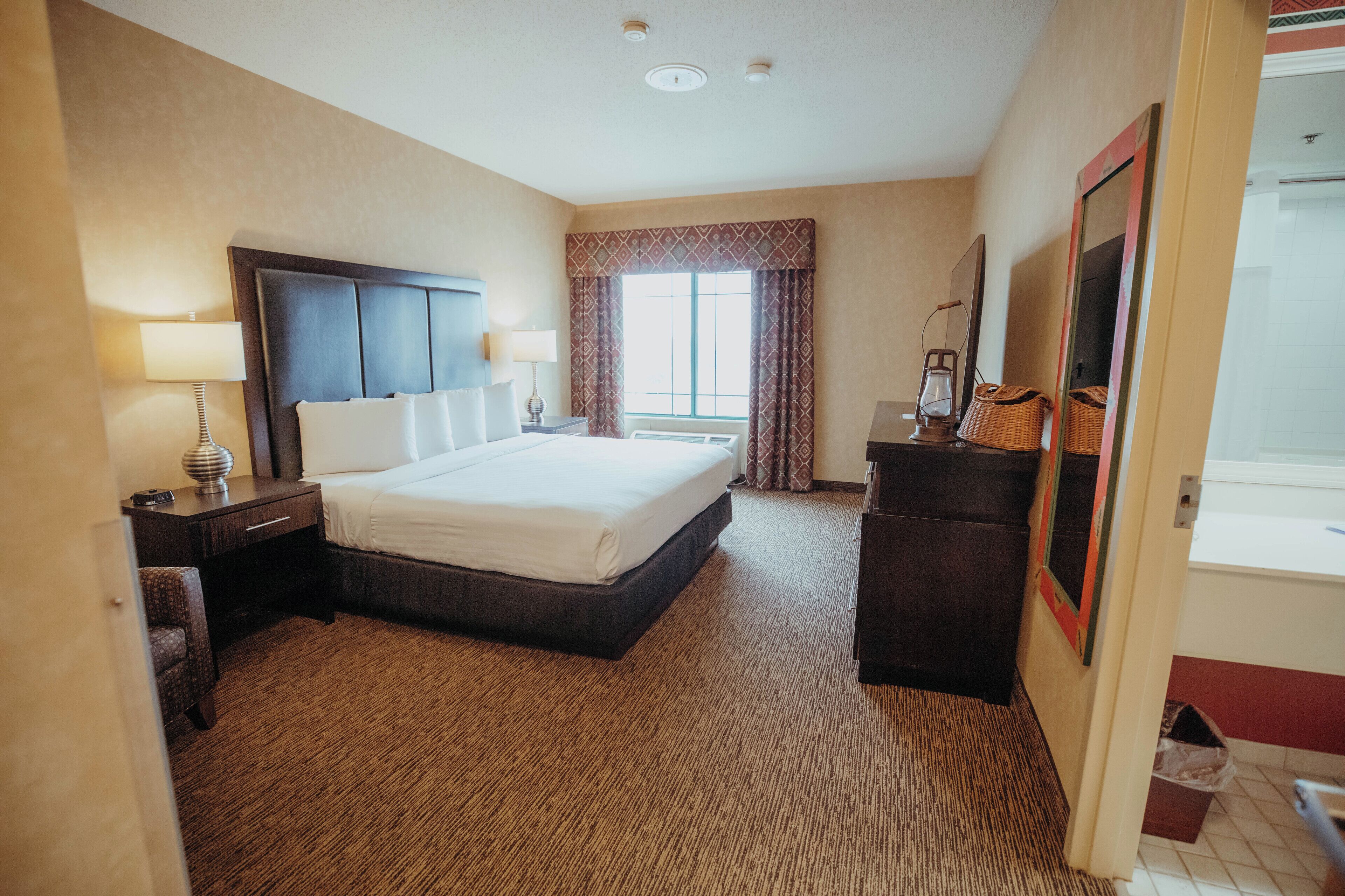 A hotel room with a large bed, a nightstand with a lamp, a dresser, and a window with curtains.