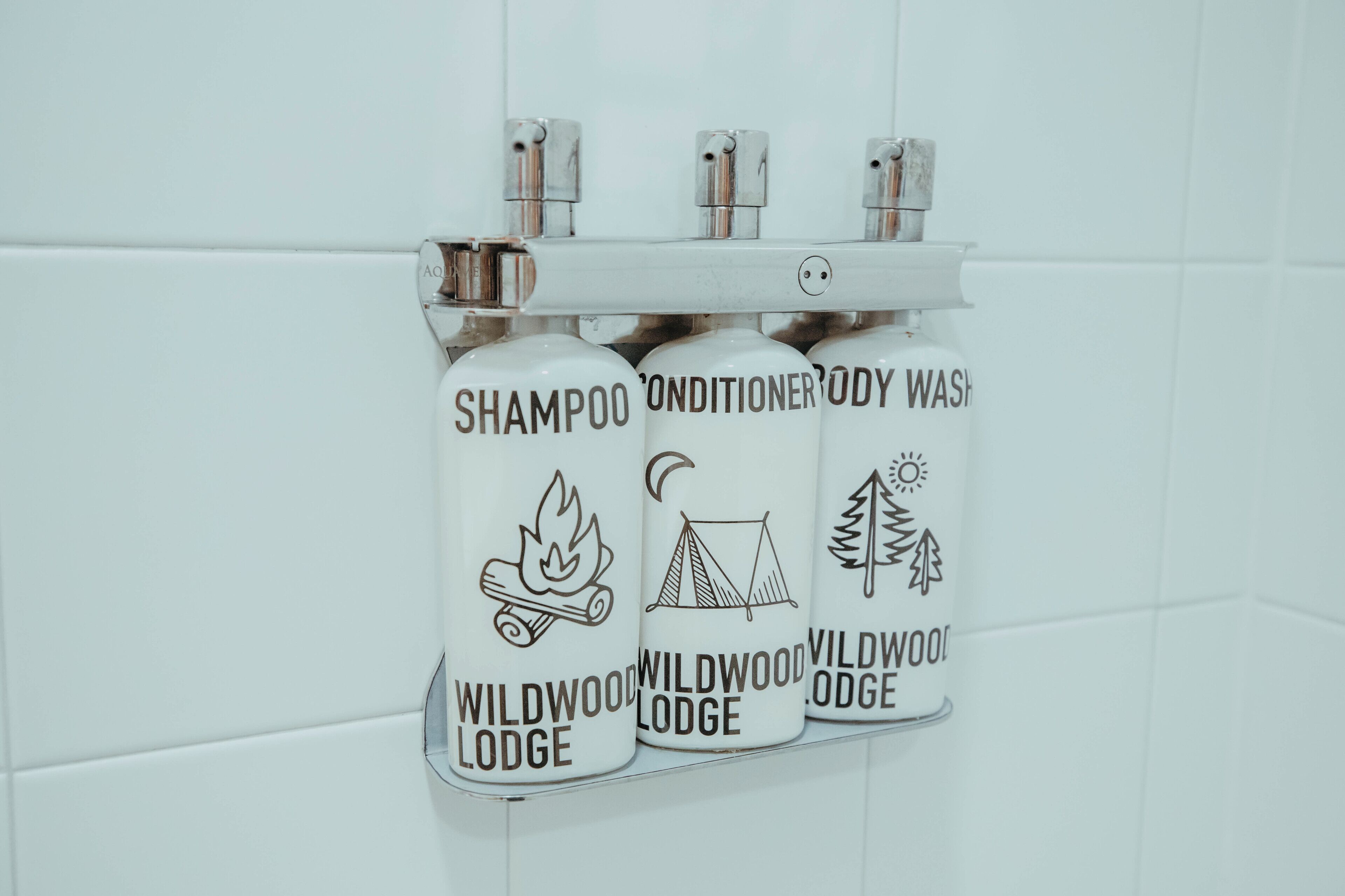 A bathroom with a white tiled wall and a mounted dispenser holding three bottles labeled 'Wildwood Lodge' with icons of a campfire, tent, and tree.