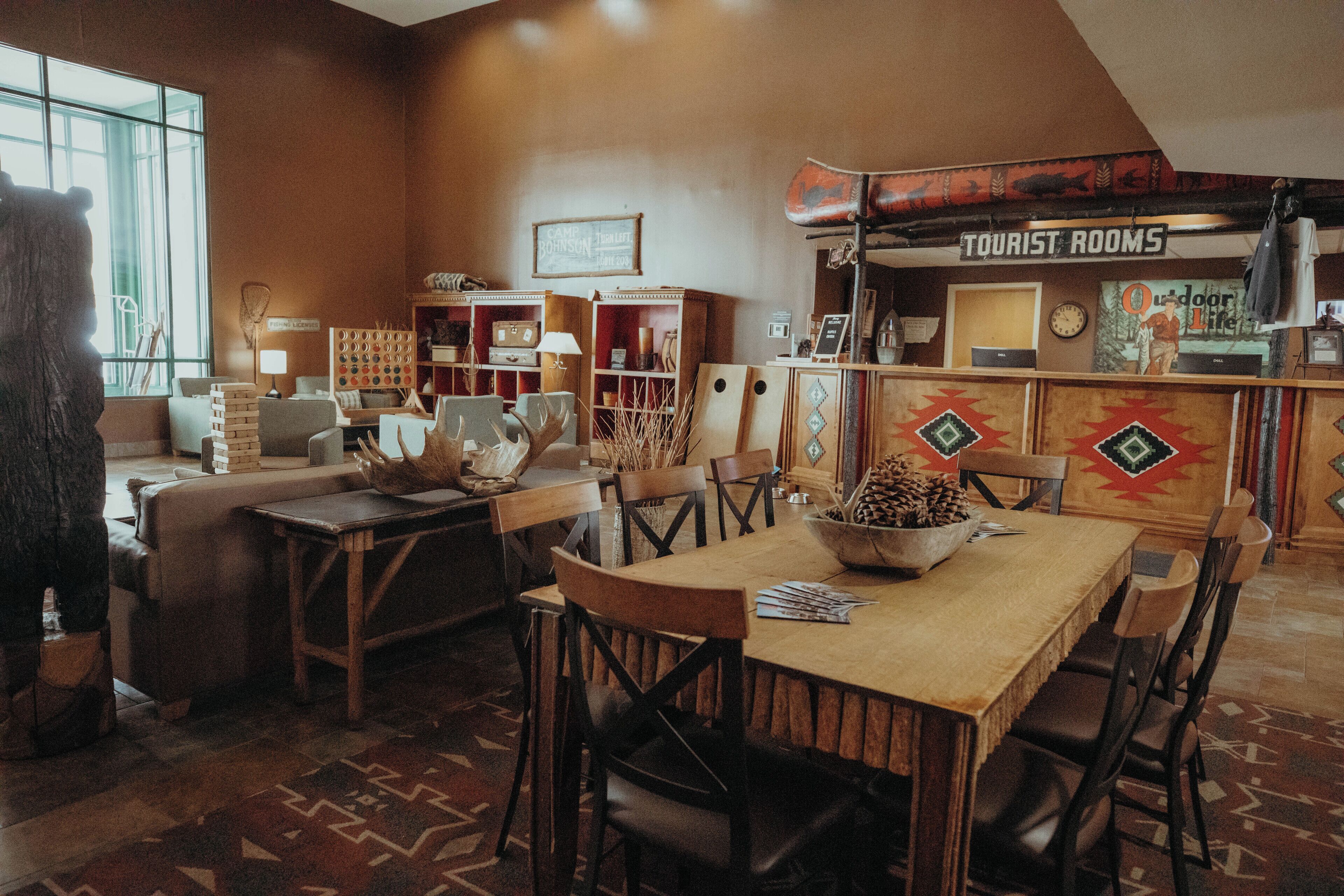 A cozy, rustic interior with a wooden dining table, chairs, and a large window. There is a sign indicating 'Tourist Rooms' and a wall-mounted clock.