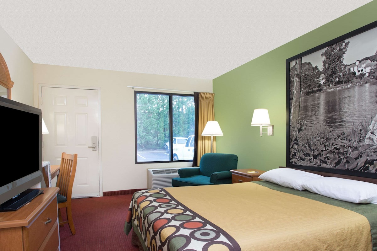 Ein Hotelzimmer mit Bett, Fernseher, Schreibtisch, Stuhl, Fenster und einem Bild an der Wand.