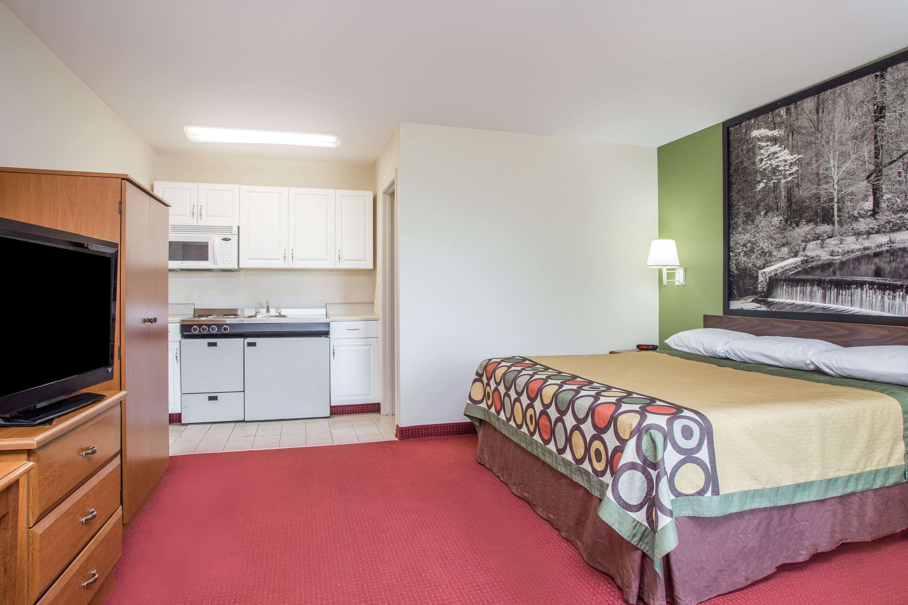 A compact hotel room with a kitchenette, a television, a bed with a patterned bedspread, and a wall-mounted lamp.