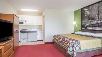 A compact hotel room with a kitchenette, a television, a bed with a patterned bedspread, and a wall-mounted lamp.