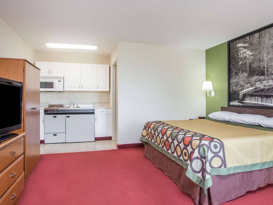 Ein kleines Hotelzimmer mit Küchenzeile, Fernseher, einem Bett mit gemustertem Überwurf und einer Wandlampe.