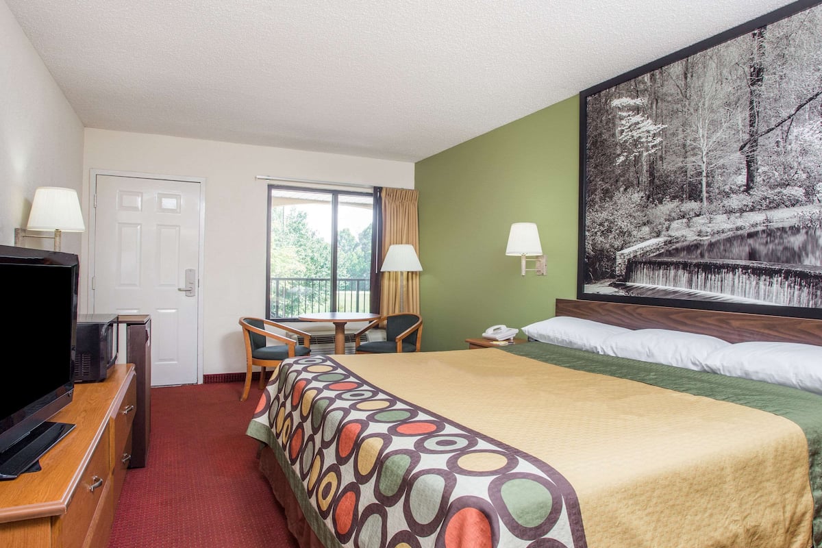 Ein Hotelzimmer mit einem Bett, einem Schreibtisch, einem Fernseher und einem Gemälde einer Waldlandschaft.
