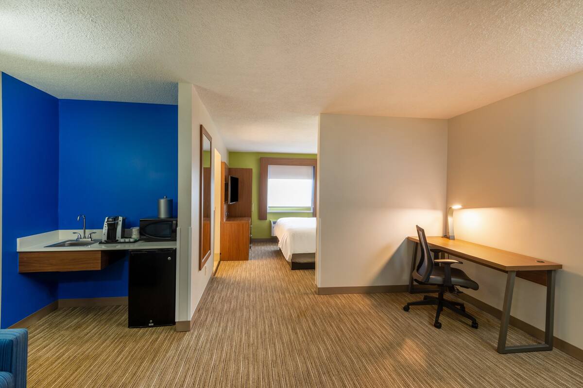 A compact hotel room with a kitchenette, a bed, a desk with a chair, and a window providing natural light.