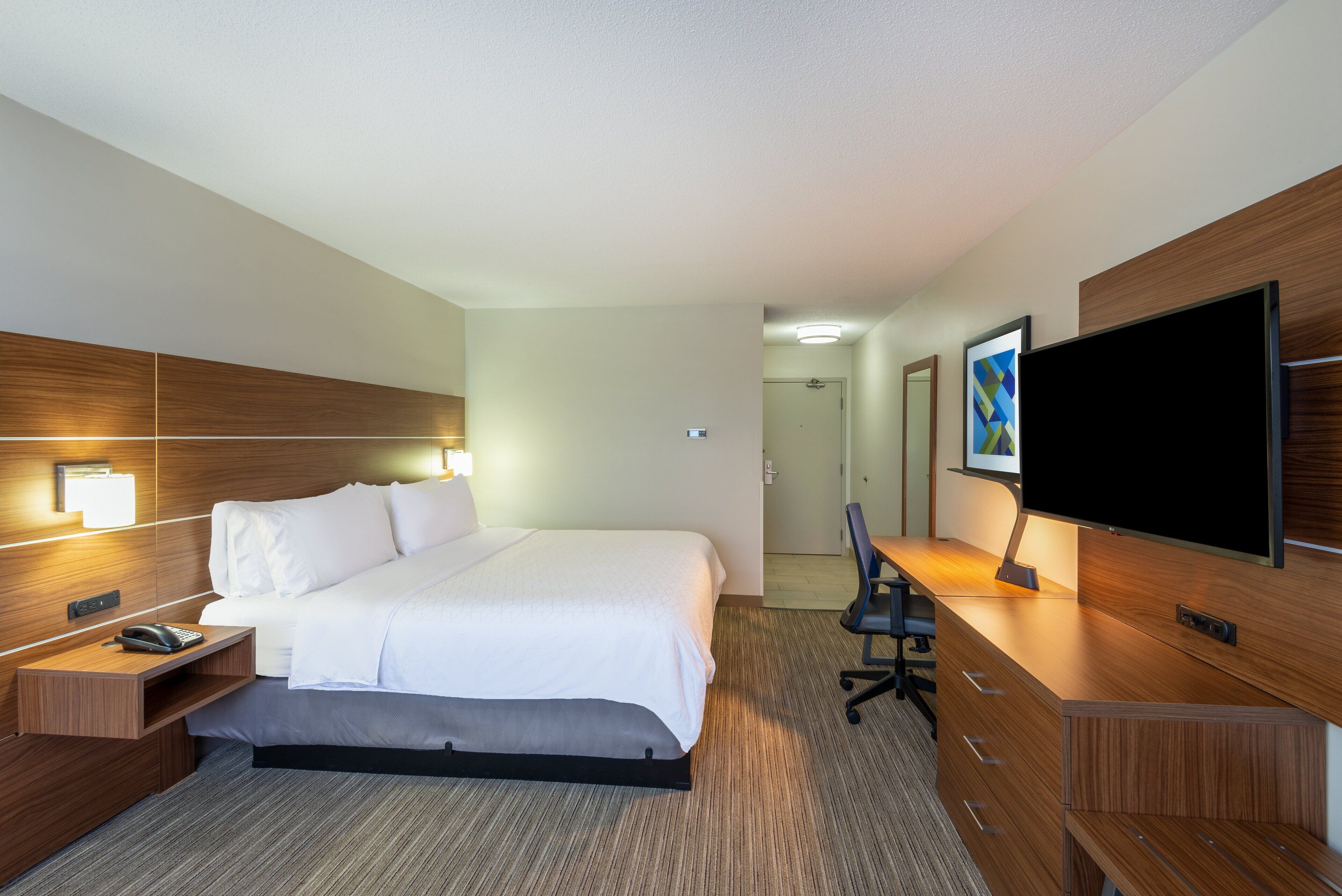 A hotel room with a bed, bedside table with a phone, a desk with a chair, a flat-screen TV, and a painting on the wall.