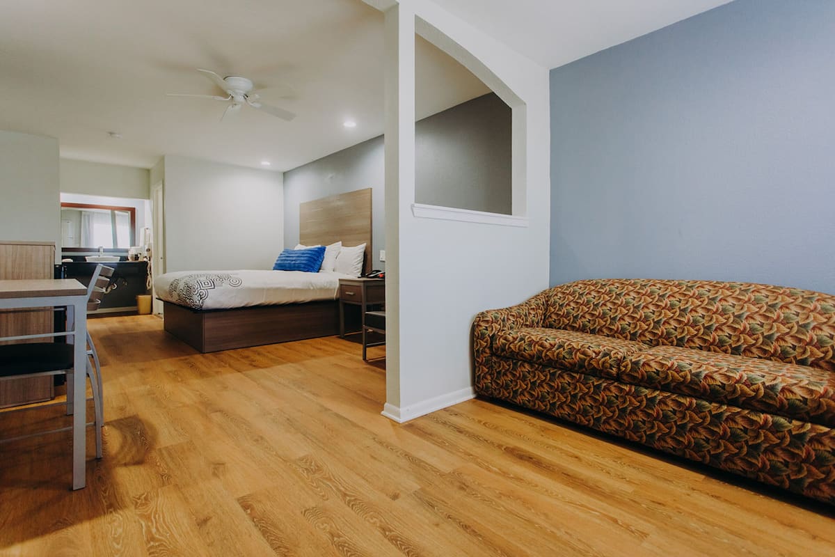 A modern hotel room with a wooden floor, a bed with patterned bedding, a sofa, a desk, and a ceiling fan.