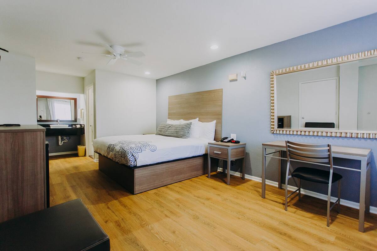 A modern hotel room with a wooden floor, a bed with a headboard, a desk with a chair, a mirror, and a ceiling fan.