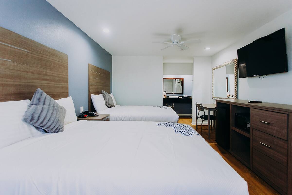 A hotel room with two beds, a desk, a chair, a television, and a ceiling fan.