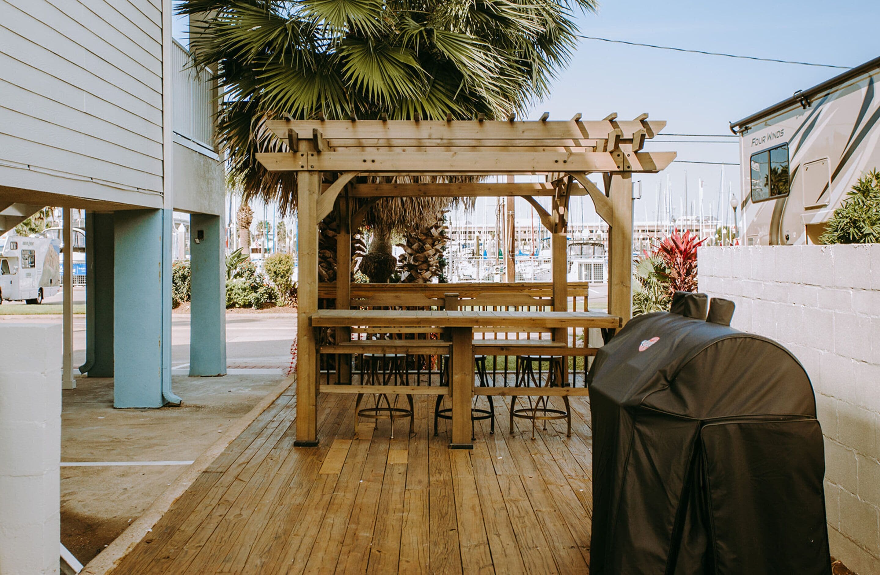 A wooden pergola with a table and chairs, a barbecue grill, and a palm tree.