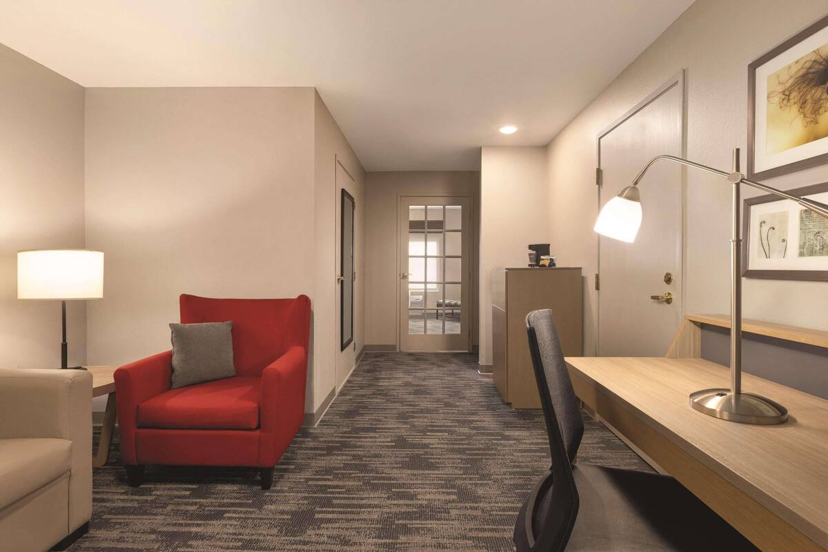 A hotel room with a red armchair, a desk with a lamp, a chair, and a carpeted floor.