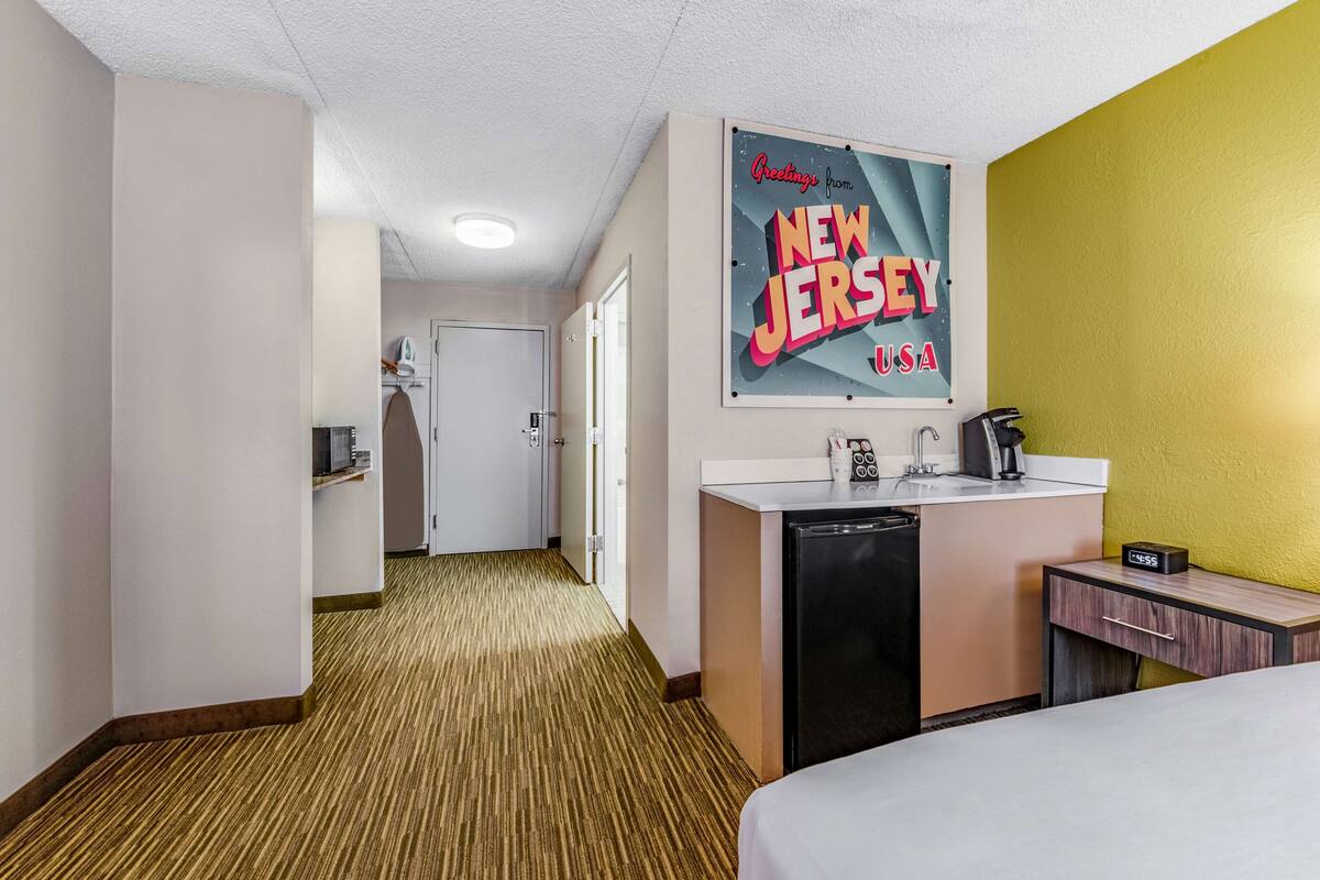 Habitación de hotel con una cama, un escritorio pequeño con lavabo, un espejo y un cartel en la pared.