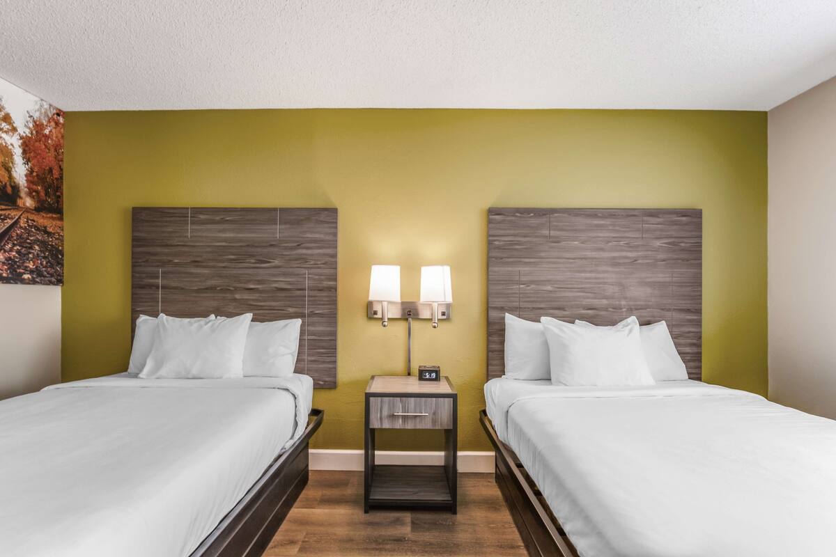 Habitación de hotel con dos camas, una mesita de noche con un reloj y una lámpara de pared.