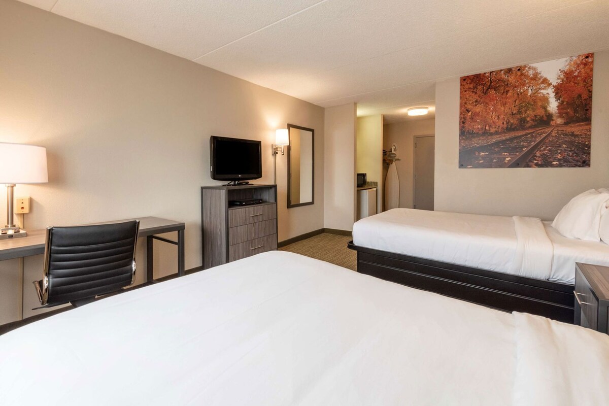 Habitación de hotel con dos camas, televisión, escritorio con lámpara, silla y un cuadro de un tren.