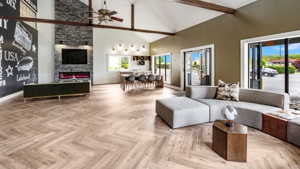 A spacious living area with a herringbone wood floor, a large sofa, a coffee table, and a fireplace. There is a dining area with a table and chairs, and a kitchen in the background. - SureStay Plus Hotel by Best Western McGuire AFB Jackson (Cookstown)
