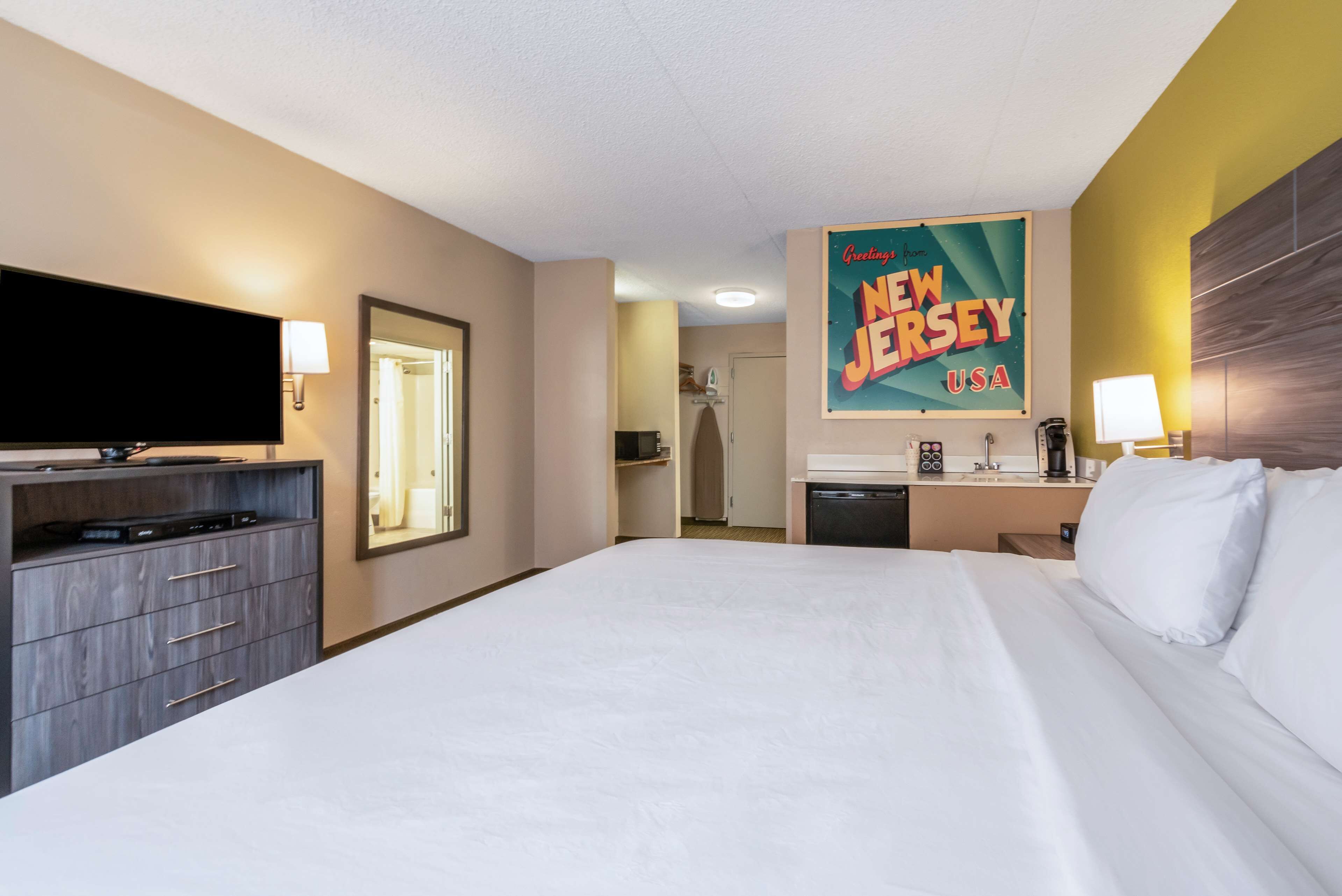 A hotel room with a large bed, a flat-screen TV, a desk with a lamp, and a wall-mounted sign that reads "Greetings from New Jersey, USA."