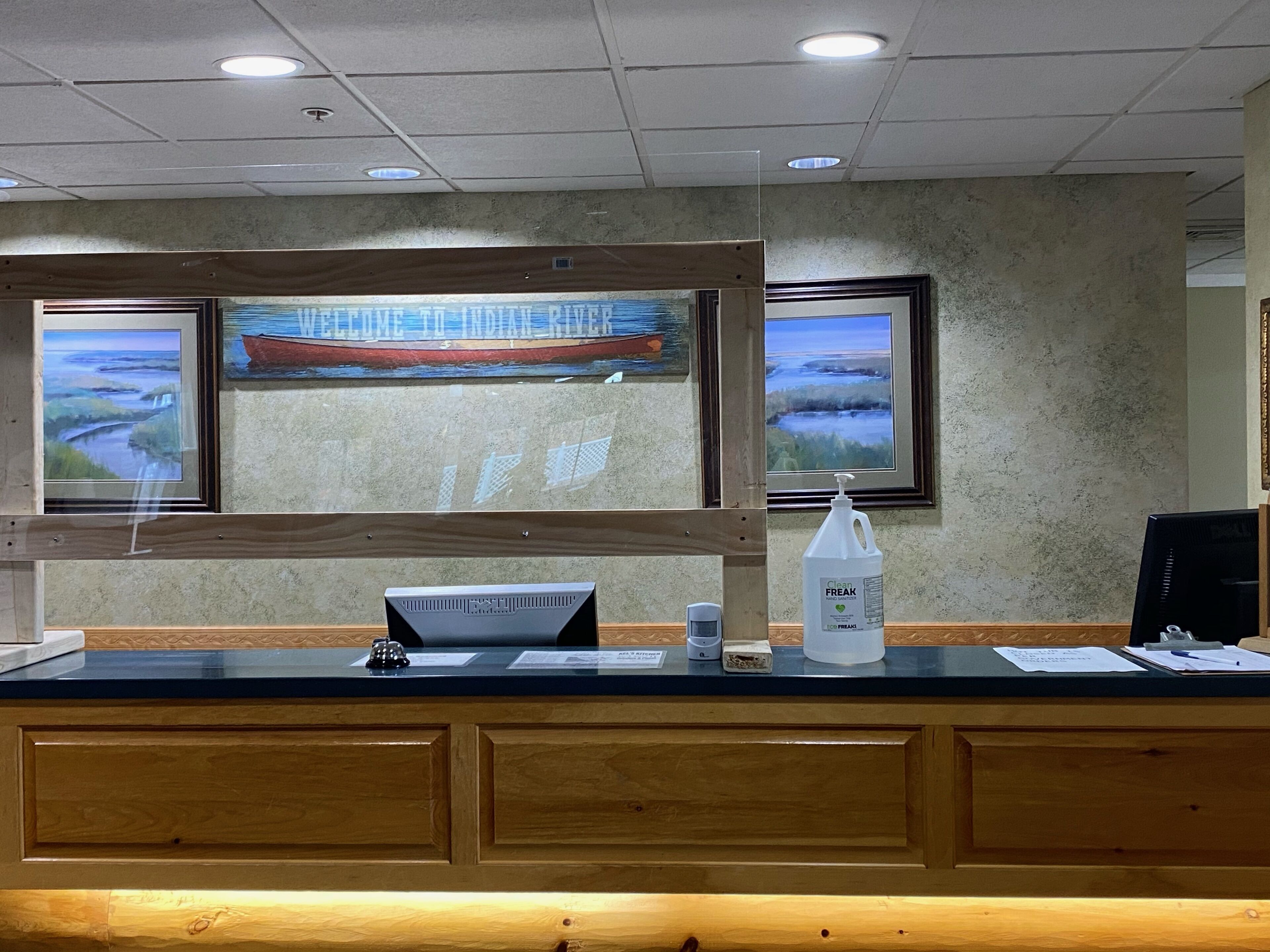 A reception desk with a wooden panel design, two framed paintings of a river scene, and a bottle of hand sanitizer.