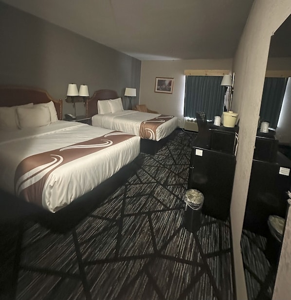 A hotel room with two beds, a desk, a chair, a trash bin, and a window with curtains. - Hometown Inn (Indian River)