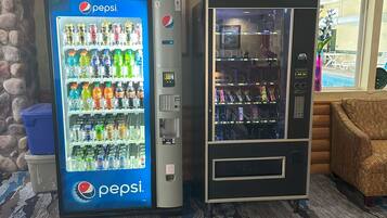 Zwei Getränkeautomaten, einer mit Pepsi und der andere mit Coca-Cola, in einem Raum mit einem gemusterten Teppich und Blick auf einen Pool.