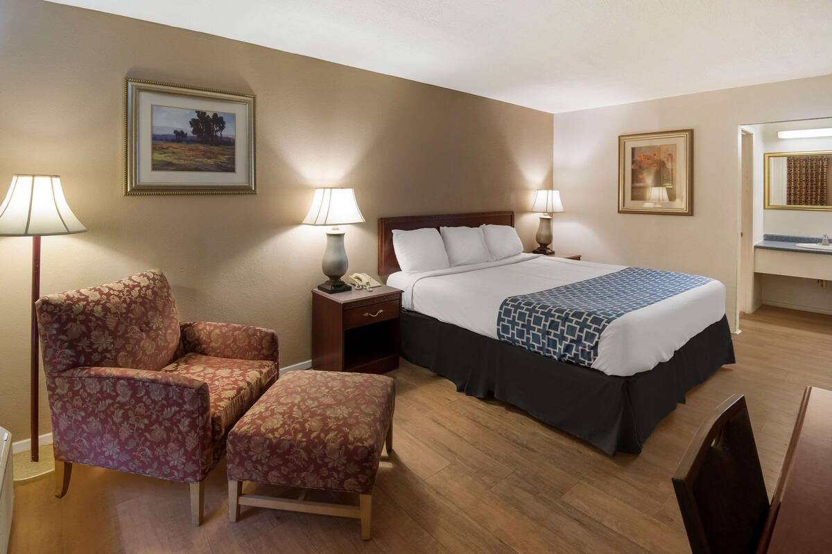 Una habitación de hotel con una cama, dos lámparas de noche, una silla, un reposapiés y una mesita de noche con un teléfono.