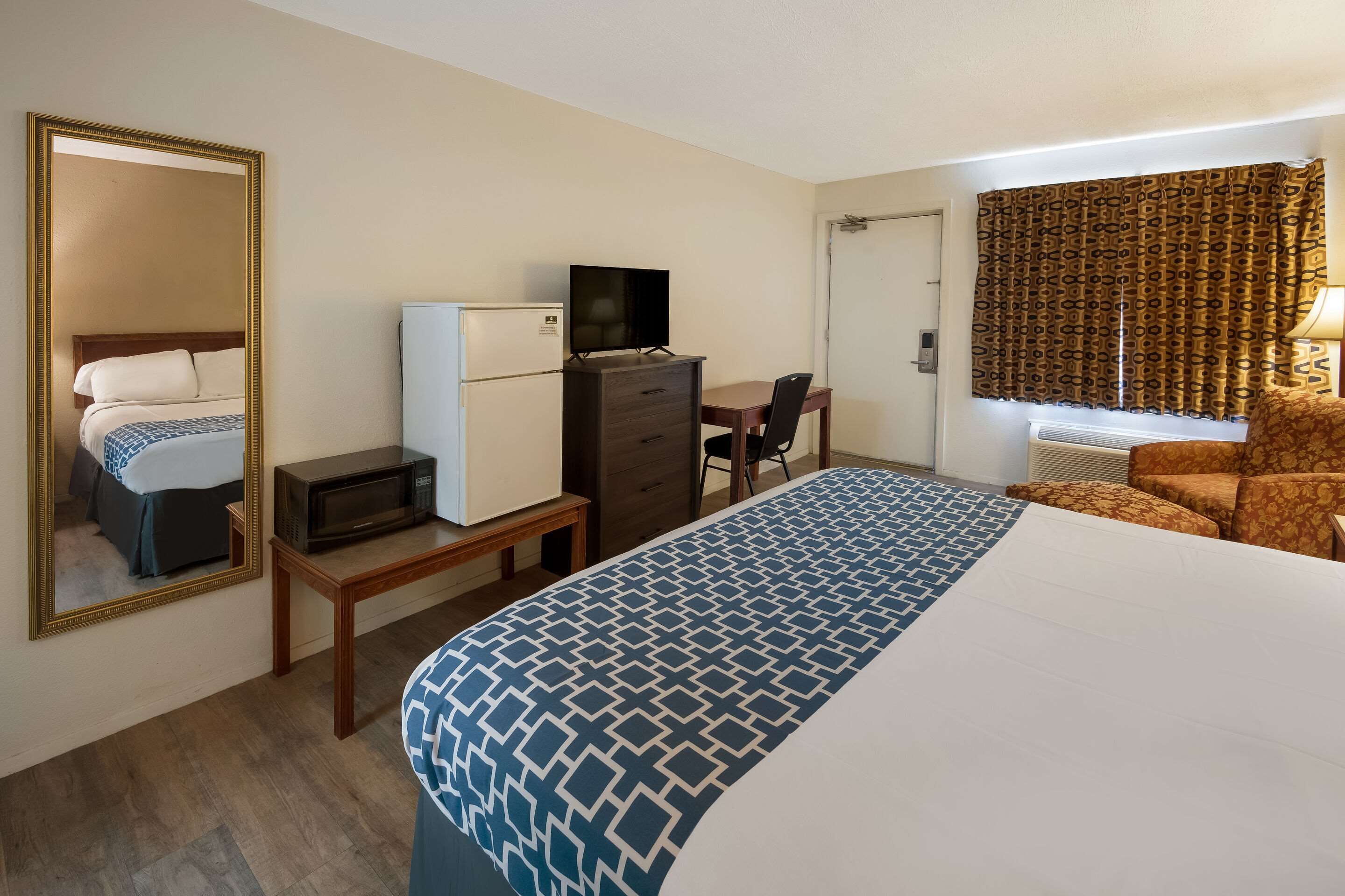 Habitación de hotel con una cama grande, un escritorio con computadora, refrigerador, microondas, una silla y un espejo amplio.
