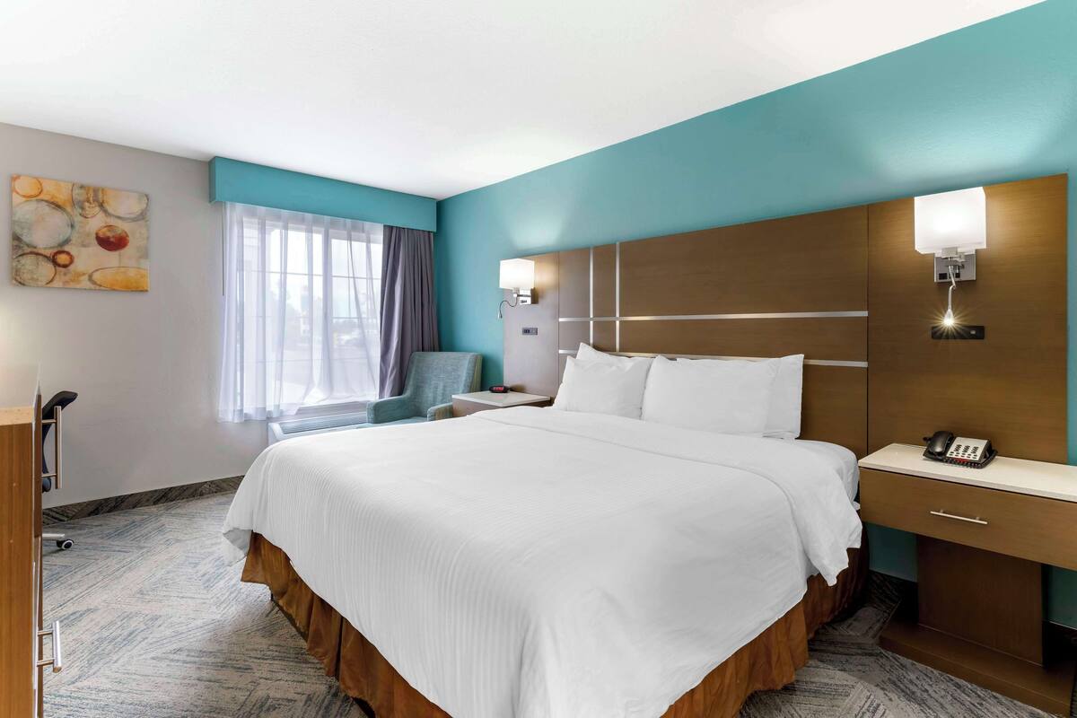 A hotel room with a large bed, a desk with a telephone, a chair, a window with curtains, and a wall-mounted light fixture.