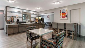 Uma sala de jantar moderna com uma mesa central e cadeiras, área de cozinha com bancada e eletrodomésticos, e parede com arte abstrata.