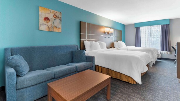 Quarto de hotel com sofá azul, mesa de madeira, cama grande com roupa de cama branca, cabeceira com luminária e pintura na parede.