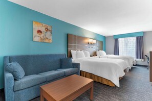 Une chambre d’hôtel avec un canapé bleu, une table en bois, un grand lit avec une literie blanche, une tête de lit dotée d’un luminaire et un tableau accroché au mur.