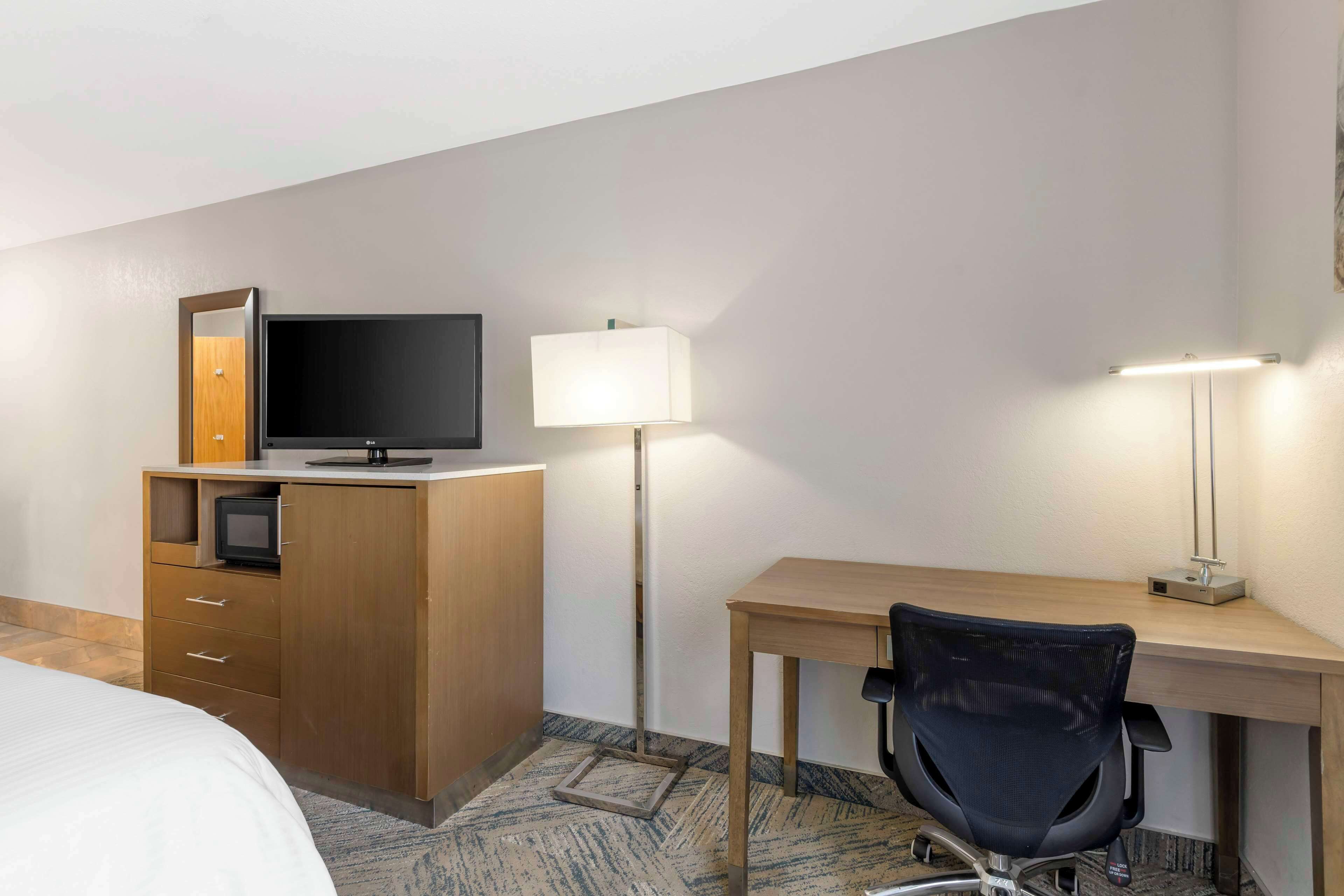 A hotel room with a bed, a desk with a chair, a television, a lamp, and a wardrobe.