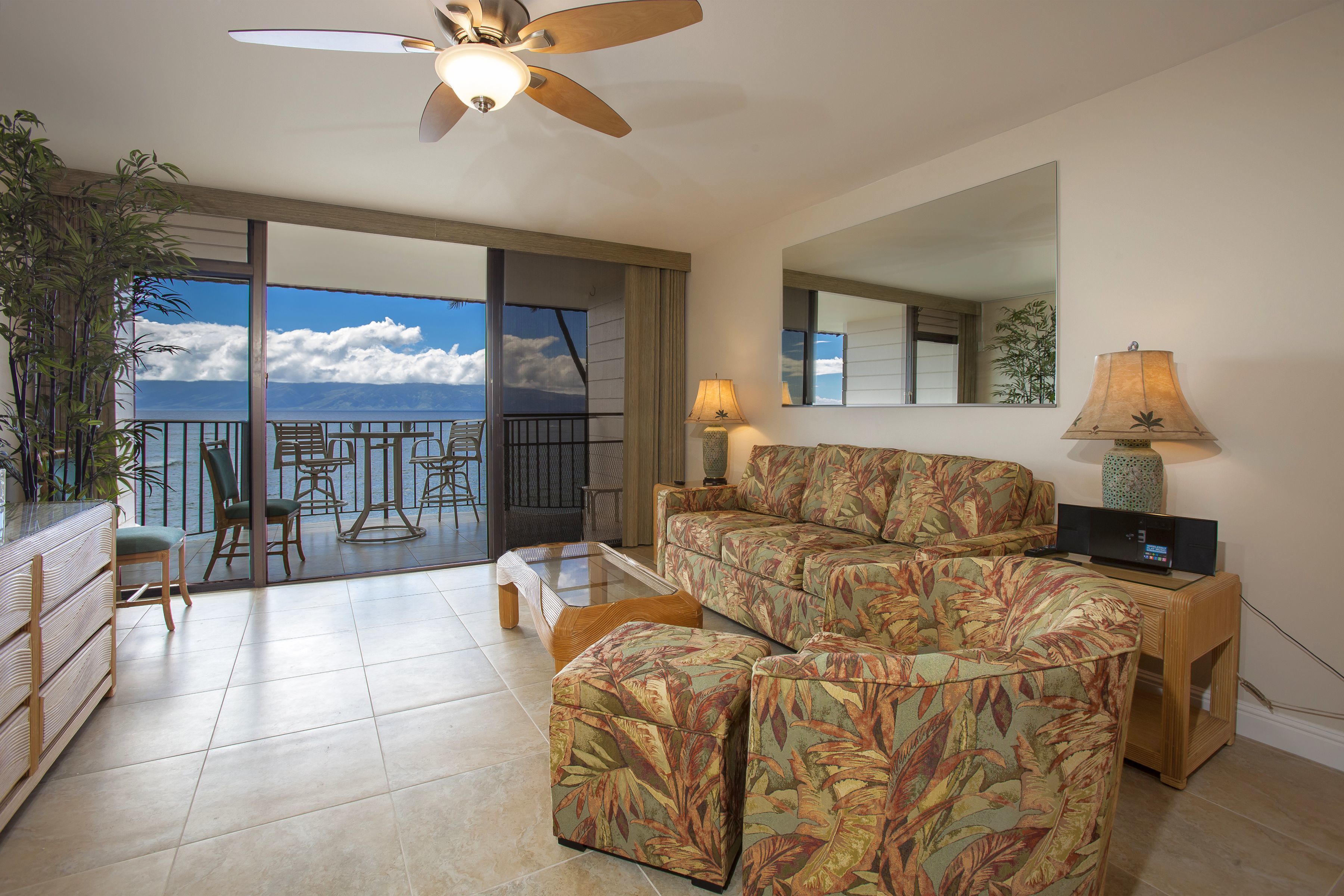 A living room with a sofa, ottoman, and a ceiling fan. There is a balcony with a view of the ocean.