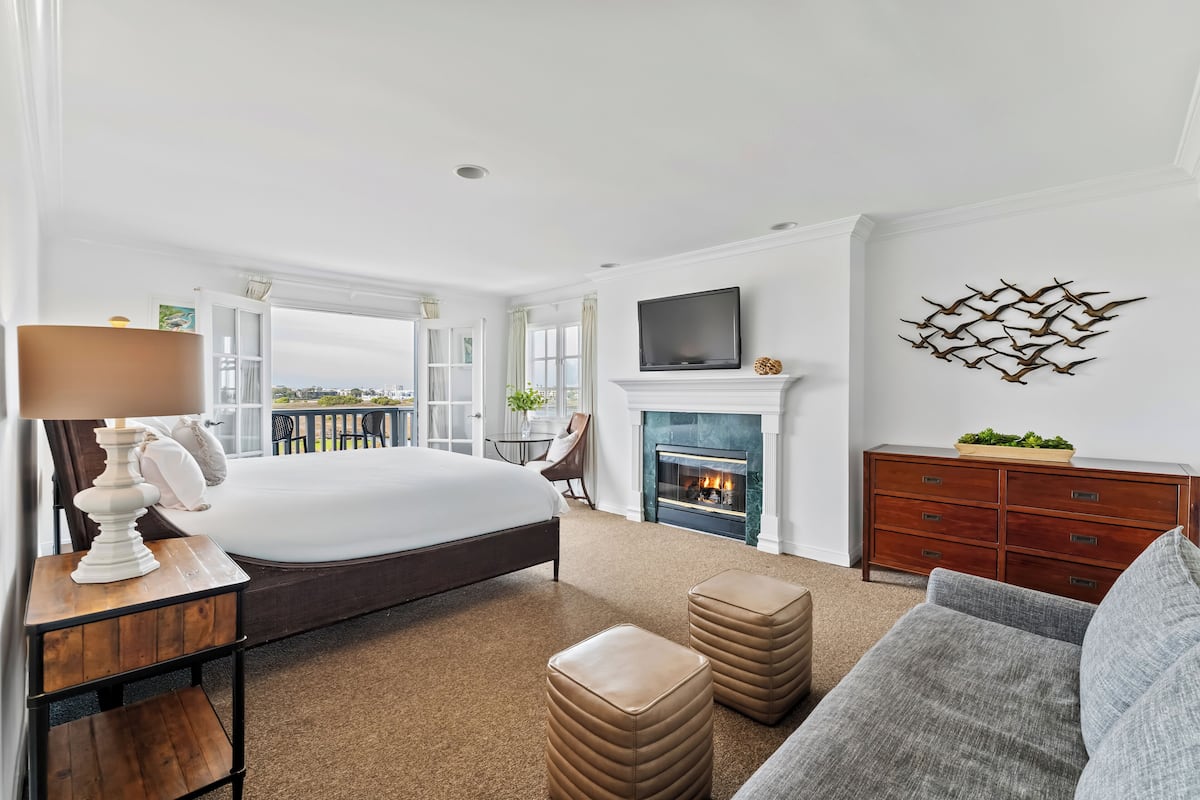 A spacious bedroom with a large bed, a fireplace, a TV, and a balcony with a view.