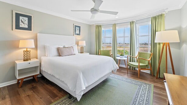 A hotel room with a large bed, a ceiling fan, a small table with a chair, and a view of the outdoors through the windows. - Inn at Playa del Rey (Playa Del Rey)