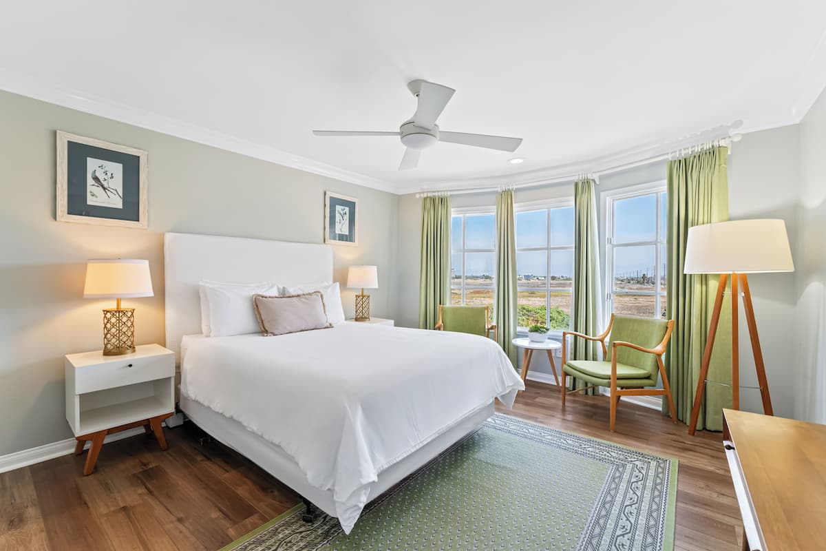 A hotel room with a large bed, a ceiling fan, a small table with a chair, and a view of the outdoors through the windows.