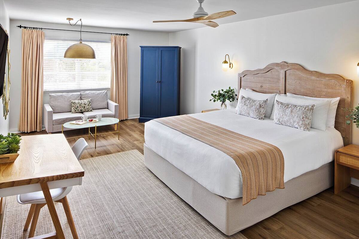 A bedroom with a large bed, a wooden headboard, a ceiling fan, a sofa, a dining table, and a window with curtains.