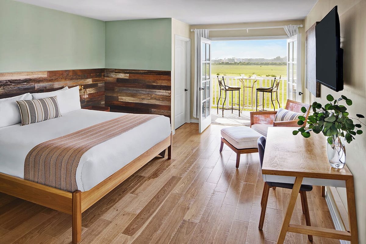 A bedroom with a large bed, a wooden headboard, a balcony with a view of the countryside, and a TV mounted on the wall.