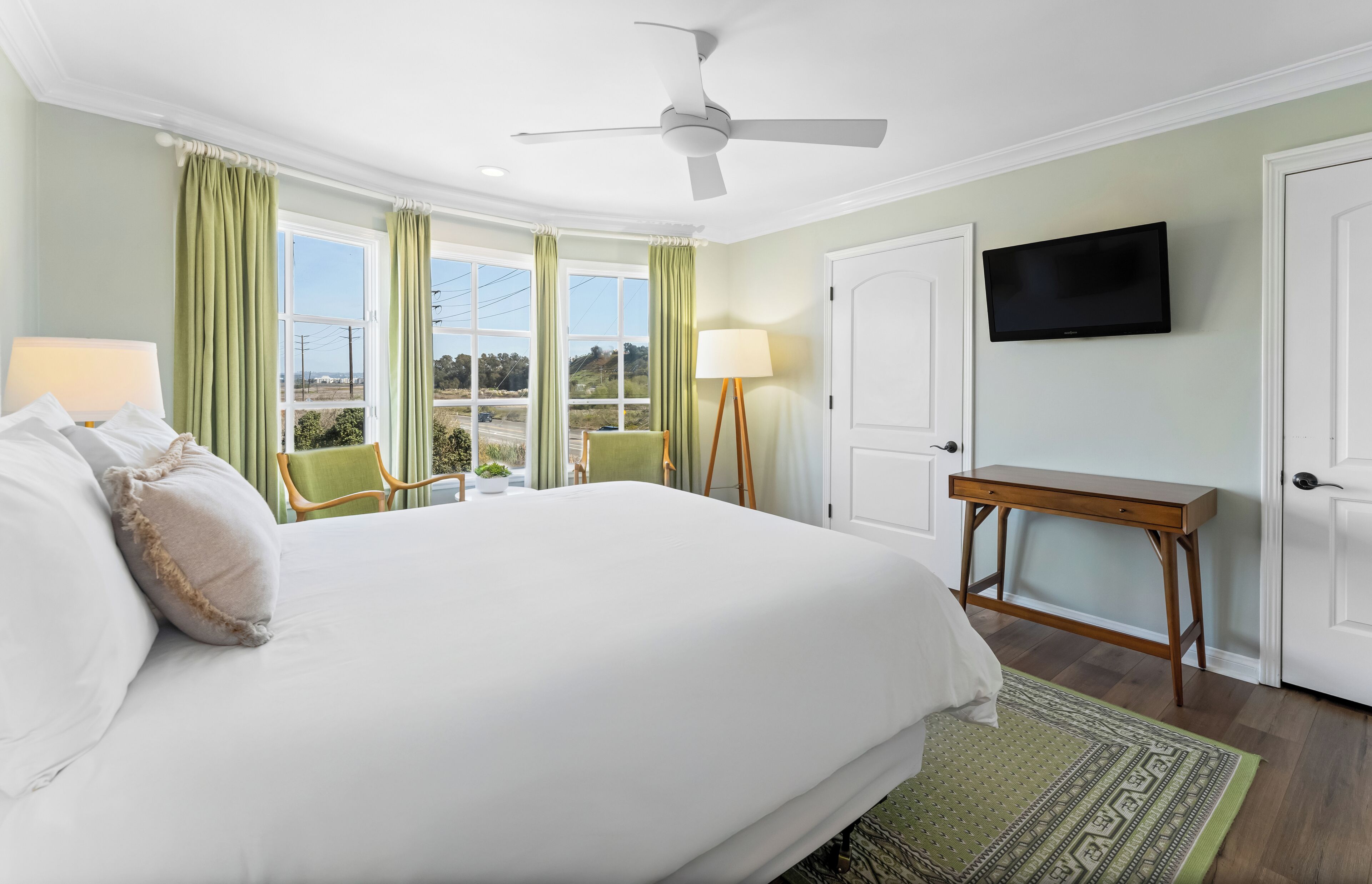 A hotel room with a large bed, a ceiling fan, a TV, a desk, and a view of the outdoors.