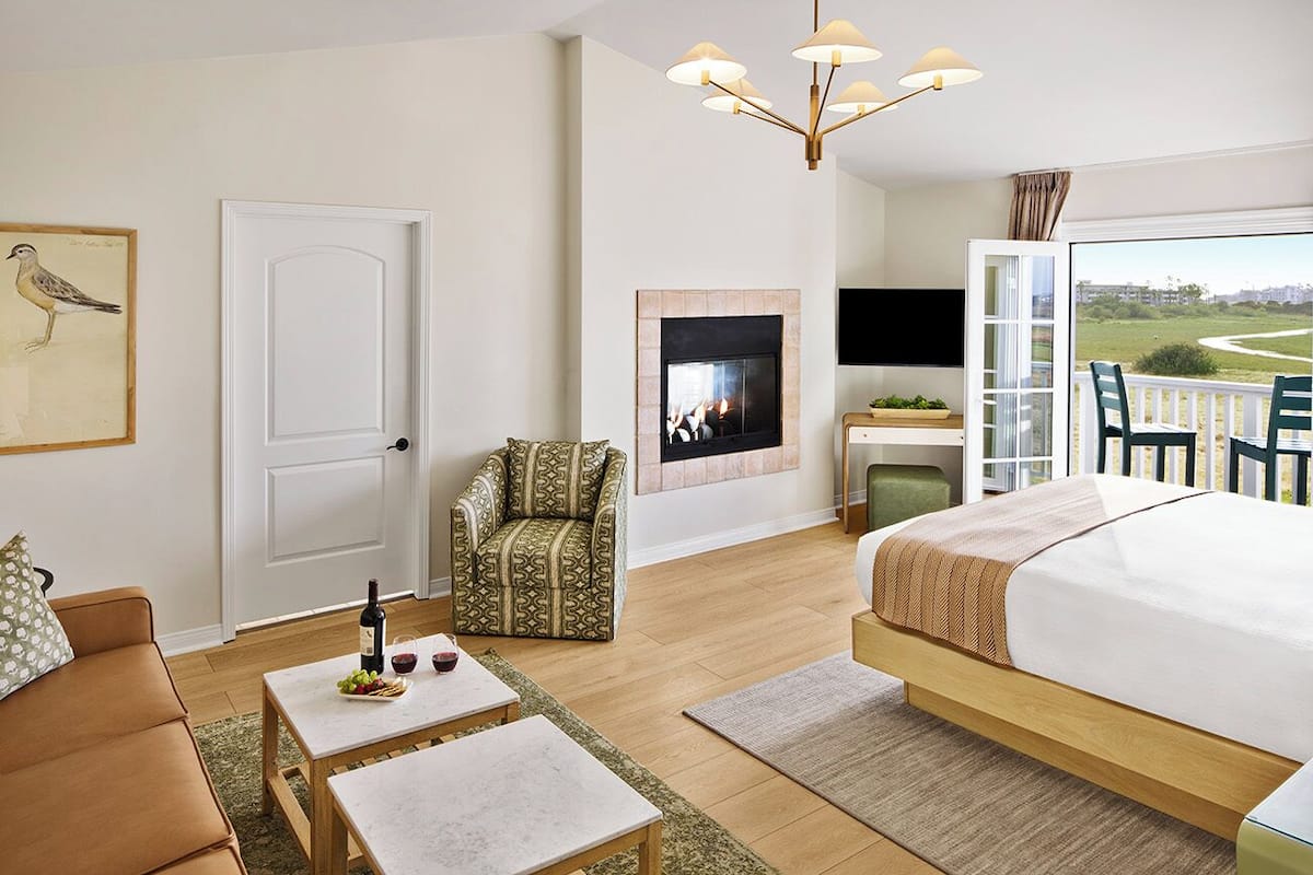 A hotel room with a bed, a sofa, a small table with a bottle and glasses, a patterned armchair, a fireplace, a TV, and a balcony with chairs.