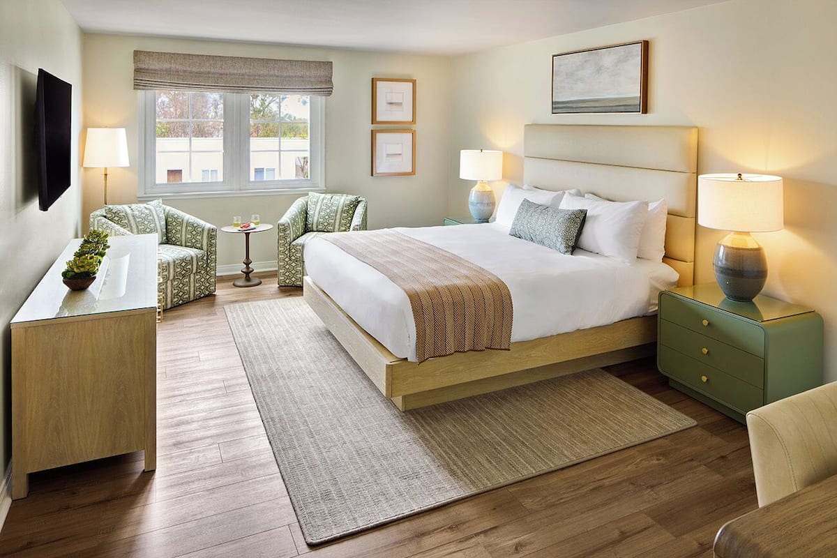 A hotel room with a large bed, bedside lamps, a desk, a chair, and a view of the outdoors through the window.